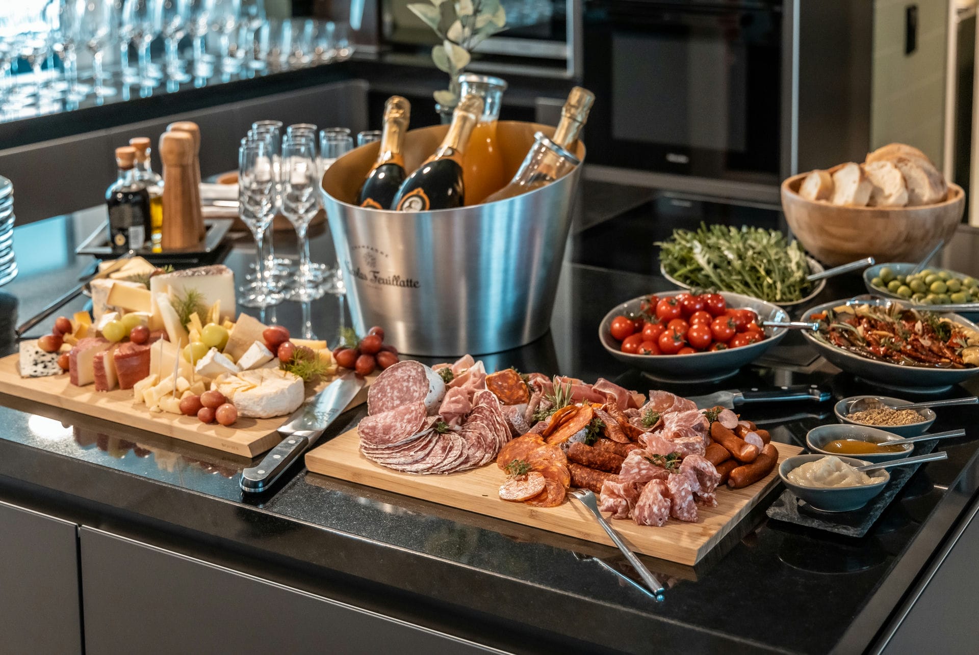 Mediterranes Buffet, Käse und Schickenauswahl auf einem großen Holzbrett, daneben Cocktailtomaten, Brot und ein Weinkühler mit Getränken.